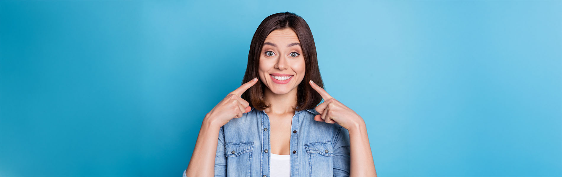 The image is a photograph of a woman with light skin, smiling at the camera. She appears to be in her late twenties or early thirties and has long hair. Her eyes are looking directly at the camera, and she is holding up her index finger near her mouth as if she s making a point or emphasizing something. The background is plain and light-colored, which suggests that this could be a stock photo used for various purposes such as advertising, personal branding, or lifestyle content.